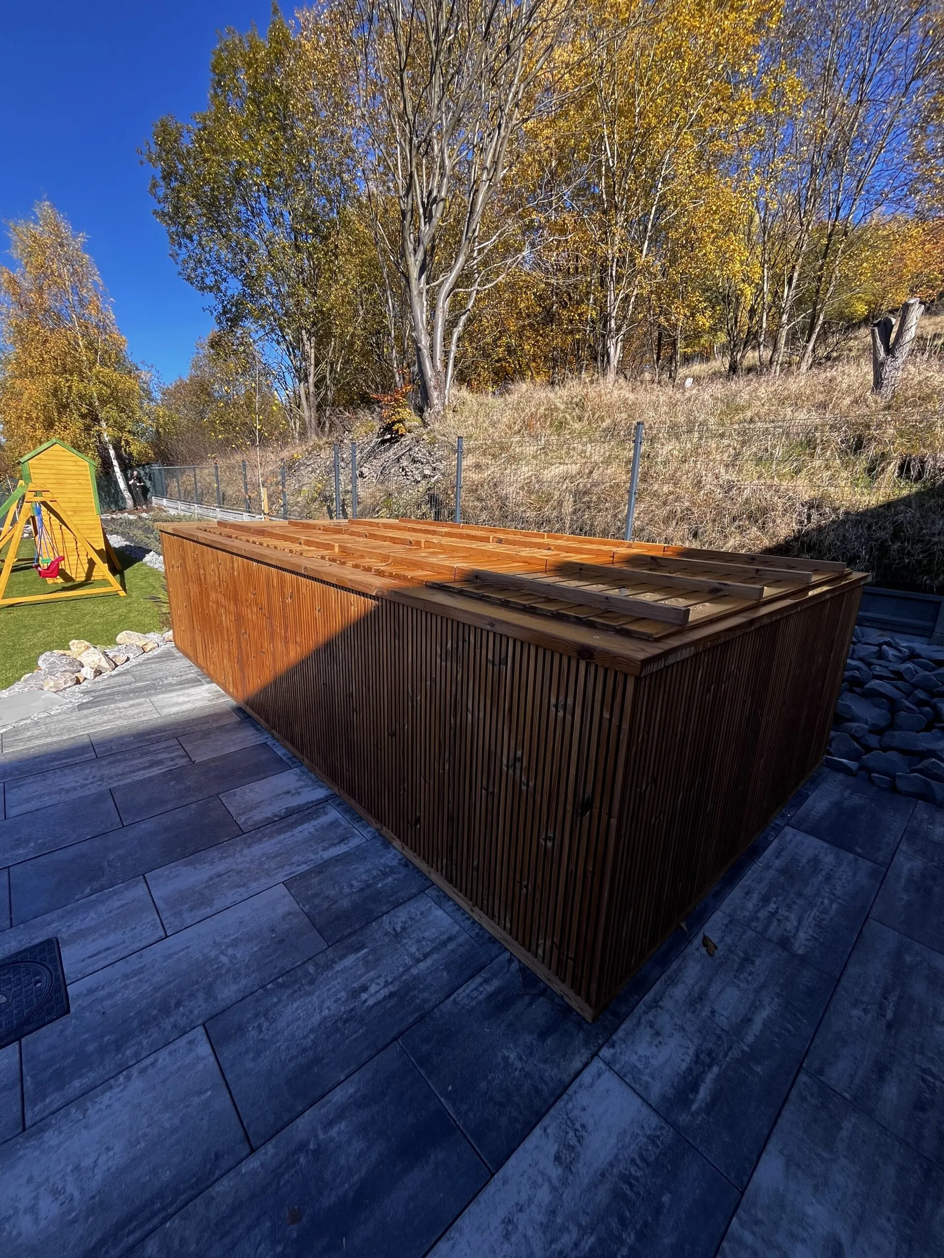 Holzpool mit Abdeckung auf einer Terrasse mit Fliesen, umgeben von herbstlichen Bäumen